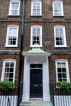 A traditional brick townhouse with three floors and symmetrical architecture. The facade features multiple white-framed windows and a prominent black front door with a white portico. Iron railings and window boxes with greenery and red flowers are present.