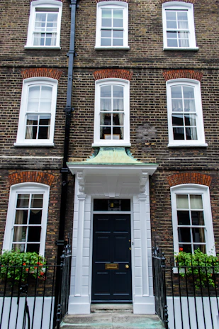 A traditional brick townhouse with three floors and symmetrical architecture. The facade features multiple white-framed windows and a prominent black front door with a white portico. Iron railings and window boxes with greenery and red flowers are present.