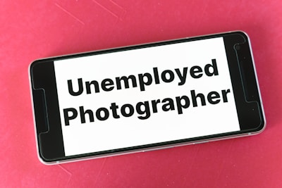 A smartphone lies on a red surface, displaying the text 'Unemployed Photographer' on its screen.