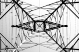 A symmetrical view from below of the intricate lattice structure of a metal electricity pylon. The geometric patterns of intersecting beams and wires create a complex web of lines against a bright sky, emphasizing the engineering and architectural design.