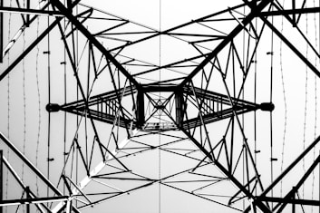 A symmetrical view from below of the intricate lattice structure of a metal electricity pylon. The geometric patterns of intersecting beams and wires create a complex web of lines against a bright sky, emphasizing the engineering and architectural design.