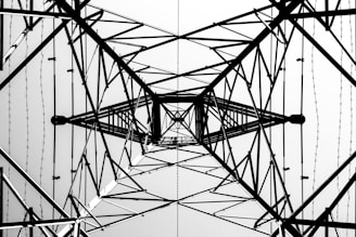 A symmetrical view from below of the intricate lattice structure of a metal electricity pylon. The geometric patterns of intersecting beams and wires create a complex web of lines against a bright sky, emphasizing the engineering and architectural design.