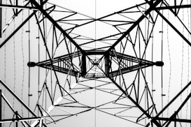 A symmetrical view from below of the intricate lattice structure of a metal electricity pylon. The geometric patterns of intersecting beams and wires create a complex web of lines against a bright sky, emphasizing the engineering and architectural design.