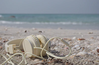 Wireless earbuds resting on a sandy shore with waves gently rolling in the background.