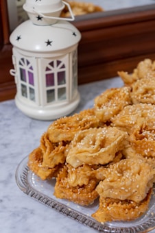 A decorative white lantern with star cutouts is placed on a marble surface next to a plate of golden-brown fried pastries sprinkled with sesame seeds. The pastries have a twisted and layered appearance.