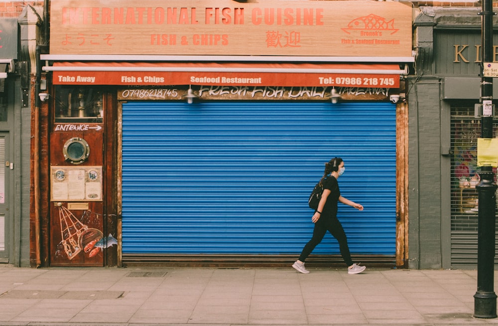 woman in black jacket and black pants standing beside blue roll up door during daytime