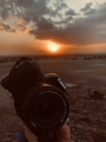 A hand is holding a Canon camera with a zoom lens, positioned against the backdrop of a serene sunset. The sun is partly hidden behind clouds, casting warm hues across the landscape.