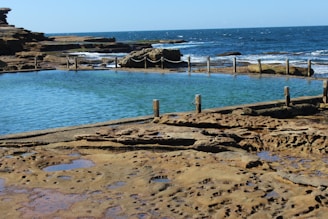 A natural rock pool with clear blue water is surrounded by a stone pathway and a rustic fence made of wooden posts and chains. Beyond the pool, waves crash against the rocky shoreline, and the ocean stretches out to the horizon under a clear blue sky.