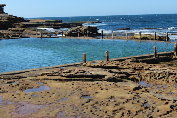 A natural rock pool with clear blue water is surrounded by a stone pathway and a rustic fence made of wooden posts and chains. Beyond the pool, waves crash against the rocky shoreline, and the ocean stretches out to the horizon under a clear blue sky.