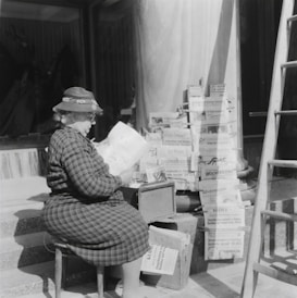An older woman sits on a stool reading a newspaper in front of a display of various newspapers arranged on racks. She wears a patterned coat and a hat adorned with flowers. The setting appears to be outdoors, with a large column and ladder to the side.