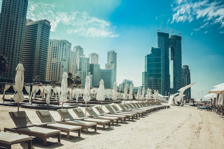 A sandy beach lined with empty lounge chairs and closed umbrellas, positioned in front of a backdrop of tall, modern skyscrapers under a clear blue sky.