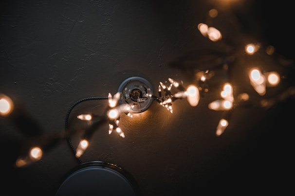 A string of warm white fairy lights is arranged in the foreground, creating a soft bokeh effect. The lights are draped around a white ceiling fixture, which appears partially in the background. The lighting is dim and atmospheric, with a focus on the glow and shape of the lights.