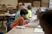 Children are seated around a table in a classroom setting, engaging in activities with paper and art supplies. The classroom appears busy with students focused and collaborating on creative tasks.