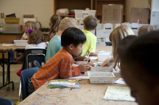Children are seated around a table in a classroom setting, engaging in activities with paper and art supplies. The classroom appears busy with students focused and collaborating on creative tasks.
