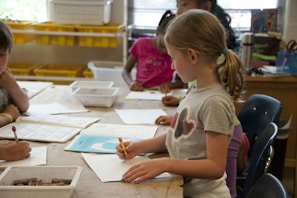Children are gathered around a table in a classroom setting, engaged in drawing and coloring activities. They have pencils and paper in front of them, with shelves of yellow storage bins in the background.