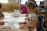 Children are gathered around a table in a classroom setting, engaged in drawing and coloring activities. They have pencils and paper in front of them, with shelves of yellow storage bins in the background.