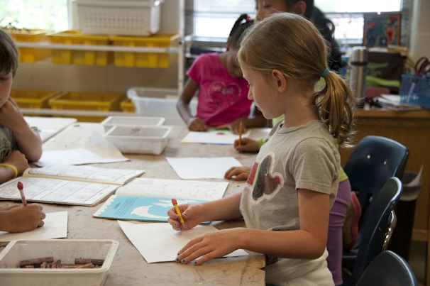 Children are gathered around a table in a classroom setting, engaged in drawing and coloring activities. They have pencils and paper in front of them, with shelves of yellow storage bins in the background.