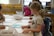 Children are gathered around a table in a classroom setting, engaged in drawing and coloring activities. They have pencils and paper in front of them, with shelves of yellow storage bins in the background.