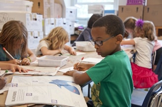 Children are seated at a table in a classroom, engaged in drawing and coloring activities. A boy in a green shirt is in focus, concentrating on his work. Other children are focused on their own projects, with art supplies scattered on the table.