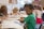 Children are seated at a table in a classroom, engaged in drawing and coloring activities. A boy in a green shirt is in focus, concentrating on his work. Other children are focused on their own projects, with art supplies scattered on the table.
