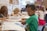Children are seated at a table in a classroom, engaged in drawing and coloring activities. A boy in a green shirt is in focus, concentrating on his work. Other children are focused on their own projects, with art supplies scattered on the table.