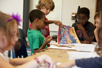 A group of children are gathered around a table engaged in an activity. One child is holding and showing a colorful book or comic to others, who are attentively looking at it. The room appears to be a classroom or a creative space with chairs and materials on the table.
