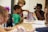 A group of children are gathered around a table engaged in an activity. One child is holding and showing a colorful book or comic to others, who are attentively looking at it. The room appears to be a classroom or a creative space with chairs and materials on the table.