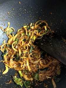 Slices of onion and green herbs are being sautéed in a pan with a rich, spicy mixture. The ingredients are coated with a dark reddish-brown seasoning, and a wooden spatula is seen stirring them.