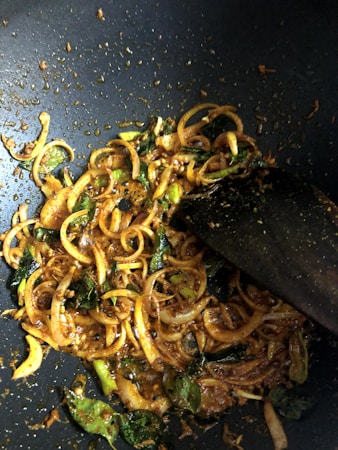 Slices of onion and green herbs are being sautéed in a pan with a rich, spicy mixture. The ingredients are coated with a dark reddish-brown seasoning, and a wooden spatula is seen stirring them.
