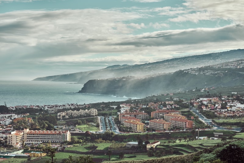 View to the district El Durazno, which belongs to Puerto de la Cruz on Tenerife. Hotel buildings and wide streets are visible. In the background is a coastal area with steep cliffs and stormy sea.