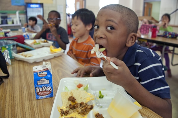 Several young children are seated at a cafeteria table enjoying their lunch. The boy in the foreground has a fork with food in his mouth and is looking at the camera. The lunch tray in front of him contains various foods including nachos with meat and cheese, fruits and vegetables, and a carton of TruMoo chocolate milk. The background shows other children eating and enjoying their meals. The setting appears to be a school cafeteria.