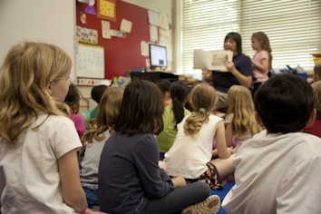 A group of young children sits on the floor of a classroom, attentively listening to a teacher reading a book. The teacher is seated in front of the children, holding up the book, while another child stands nearby. The classroom is decorated with various educational materials, and a computer is visible on a desk. Natural light filters through the blinds on the window.