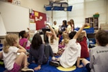 Children sit on a colorful carpet in a classroom, raising their hands eagerly towards a teacher seated in the front. The room is filled with educational decorations, bookshelves, and various classroom supplies. The atmosphere is lively and engaging as the children participate in an interactive session.