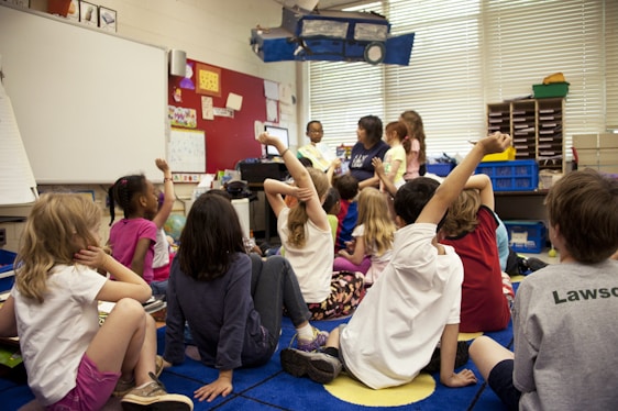 Children sit on a colorful carpet in a classroom, raising their hands eagerly towards a teacher seated in the front. The room is filled with educational decorations, bookshelves, and various classroom supplies. The atmosphere is lively and engaging as the children participate in an interactive session.