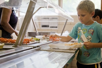 A young boy with light hair is selecting food items at a cafeteria counter. He holds a tray with different compartments and is picking up what appears to be carrots from a food station. Various vegetables are displayed on trays behind the counter, and a cafeteria worker is visible in the background.