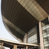 A modern architectural structure with a large, curved roof and glass windows. The building is prominently labeled 'Dewan Agung Tunku Canselor'. The facade showcases geometric shapes and a combination of concrete and glass elements, conveying an innovative design.
