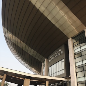 A modern architectural structure with a large, curved roof and glass windows. The building is prominently labeled 'Dewan Agung Tunku Canselor'. The facade showcases geometric shapes and a combination of concrete and glass elements, conveying an innovative design.