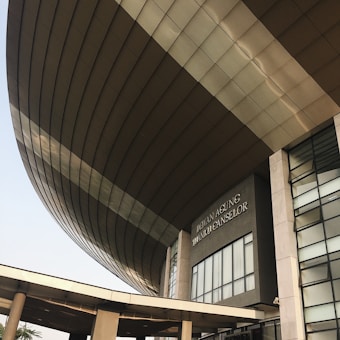 A modern architectural structure with a large, curved roof and glass windows. The building is prominently labeled 'Dewan Agung Tunku Canselor'. The facade showcases geometric shapes and a combination of concrete and glass elements, conveying an innovative design.