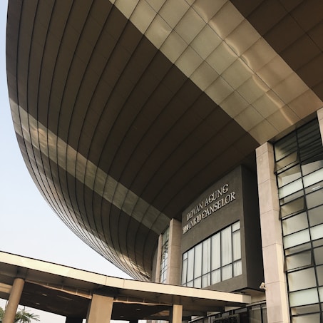 A modern architectural structure with a large, curved roof and glass windows. The building is prominently labeled 'Dewan Agung Tunku Canselor'. The facade showcases geometric shapes and a combination of concrete and glass elements, conveying an innovative design.