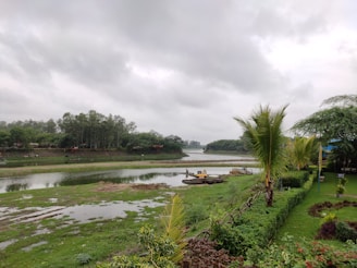 A serene landscape featuring a river surrounded by lush greenery and trees under a cloudy sky. A neat garden with trimmed hedges and palm trees borders the foreground. A small yellow excavator is visible near the riverbank.