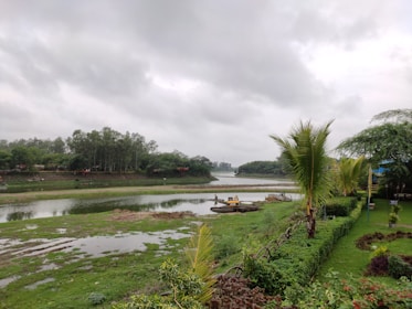 A serene landscape featuring a river surrounded by lush greenery and trees under a cloudy sky. A neat garden with trimmed hedges and palm trees borders the foreground. A small yellow excavator is visible near the riverbank.