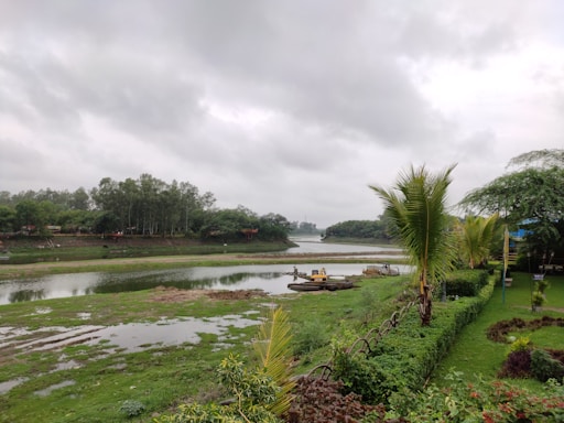 A serene landscape featuring a river surrounded by lush greenery and trees under a cloudy sky. A neat garden with trimmed hedges and palm trees borders the foreground. A small yellow excavator is visible near the riverbank.