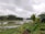 A serene landscape featuring a river surrounded by lush greenery and trees under a cloudy sky. A neat garden with trimmed hedges and palm trees borders the foreground. A small yellow excavator is visible near the riverbank.