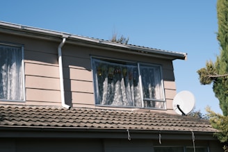 A two-story house with light beige siding and a sloped tiled roof. The upper floor windows have lace curtains, with stuffed animals visible through one window. A large satellite dish is mounted on the roof. Tall green trees are visible in the background against a clear blue sky.