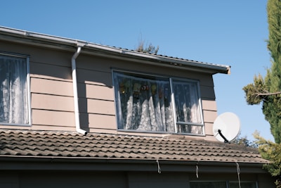 A two-story house with light beige siding and a sloped tiled roof. The upper floor windows have lace curtains, with stuffed animals visible through one window. A large satellite dish is mounted on the roof. Tall green trees are visible in the background against a clear blue sky.