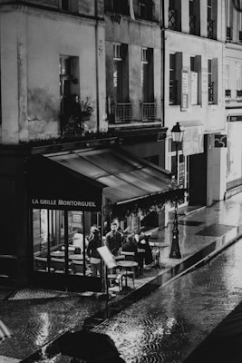A black and white photograph of a Parisian street scene captures an outdoor cafe called La Grille Montorgueil on a rainy evening. The cobblestone street glistens with reflections from streetlights and the cafe's window. People are seated under the cafe's awning, engaging in conversation and appearing relaxed despite the weather. The architecture features typical Parisian buildings with tall windows and wrought iron railings.