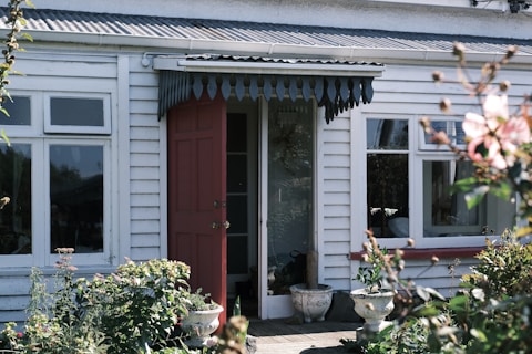 A quaint, white wooden house with a red open door stands amongst lush greenery. The front features large windows, decorative awning, and potted plants flanking the entrance. Bushes and flowers are visible in the foreground, enhancing the homely atmosphere.