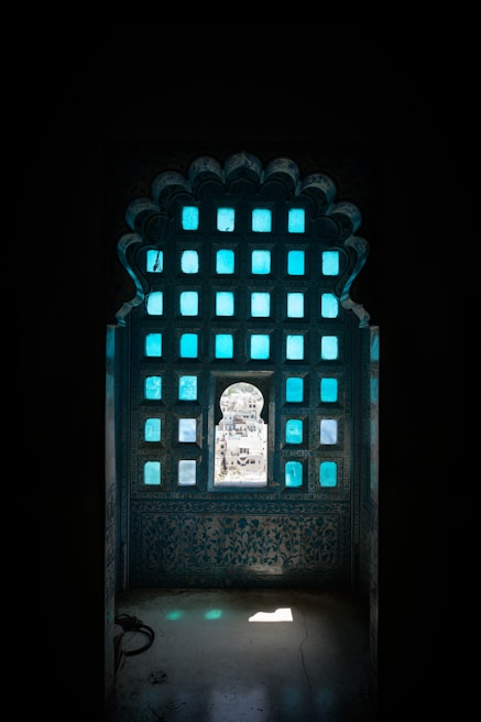 A decorative architectural window featuring a grid of blue-tinted glass panes, with ornate carving details on the surrounding stonework. Through the central arched opening, a glimpse of a bright, sunlit cityscape is visible in the background.