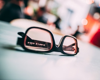 A pair of glasses with black frames resting on a light-colored surface, with text visible on the left lens. The background is blurred, adding depth and focus to the glasses.