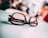 A pair of glasses with black frames resting on a light-colored surface, with text visible on the left lens. The background is blurred, adding depth and focus to the glasses.